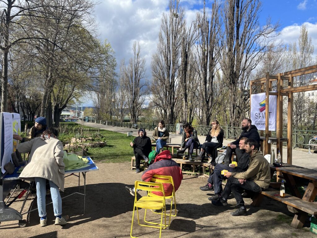 © Nicolai Benker Auf dem Bild ist der Gemeinschaftsgarten zu sehen. Rechts Sitzen Workshop Teilnehmer:innen auf einer Tribüne und Links wird auf dem Tischtennis-Tisch präsentiert