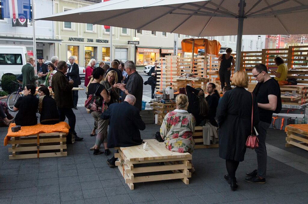 Menschen unter Schirm auf Holzinstallation sitzend und stehend