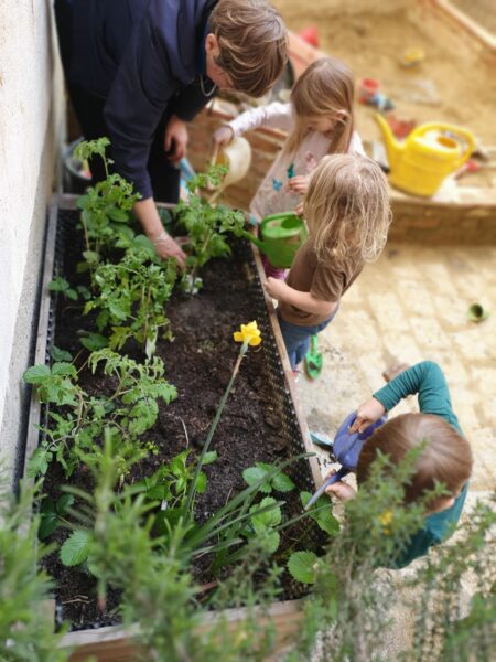 Drei Kinder und eine Erwachsene beim Gärtnern im Hochbeet. Alle drei Kinder halten Gießkannen und wässern die Pflanzen. Im Hintergrund ist eine Sandkiste mit einer weiteren Gießkanne drauf und Sandspielzeug im Hintergrund. Copyright Gerhild Soyka-Silber.