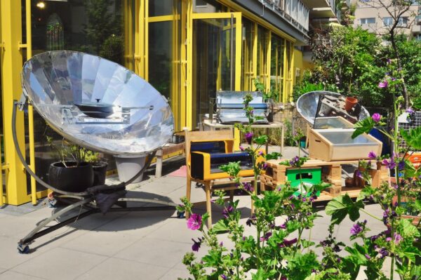 Solarmanufaktur. Eine Terrasse mit vielen Pflanzen im Sonnenlicht. Es stehen mehrere Gerätschaften herum; wie große metallische Teller, die die Sonne bündeln und mit denen dann gekocht wird. Copyright Daniel Nasr