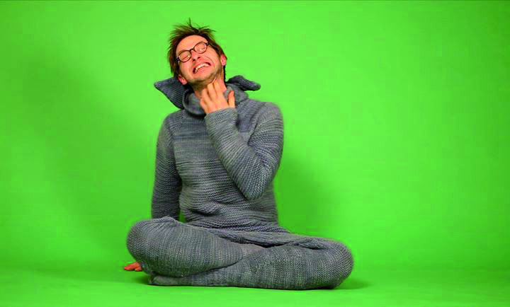 Mann mit Brille am Boden sitzend in einem grauen Onesie, grüner Hintergrund