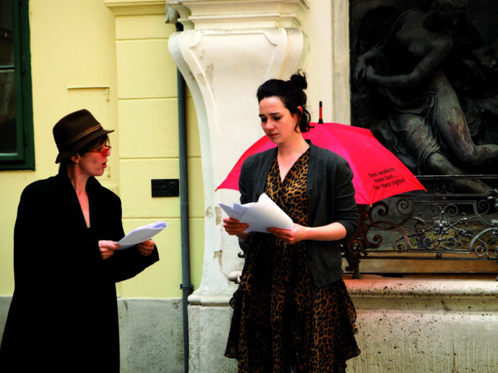 Zwei Frauen mit Zetteln in der Hand auf der Straße, dahinter ein Regenschirm mit der Aufschrift "Sex workers have lust .... for their rights!"