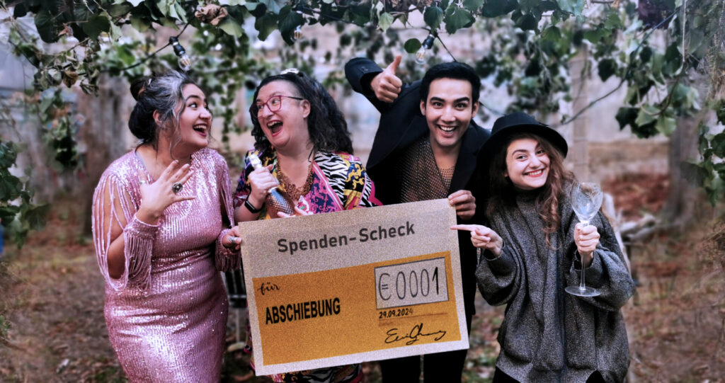 4 Personen in festlichen Outfits mit freudigem Gesichtsausdruck halten einen Spenden-Scheck auf dem steht 