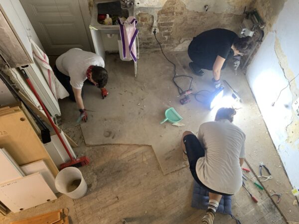 Drei Personen arbeiten gemeinsam auf engem Raum in einem renovierungsbedürftigen Raum, um den alten Parkettboden freizulegen. Die Wände sind teils verputzt, teils liegt das Mauerwerk frei. Eine helle Baustellenlampe beleuchtet den Bereich. Auf dem Boden liegen Werkzeuge wie Schraubenzieher, Zangen, ein Akkuschrauber, sowie ein kleiner Handfeger und eine Kehrschaufel. Eine Person kniet auf einem Polster, eine andere trägt Handschuhe und arbeitet in der Ecke, während die dritte in der Nähe der Tür arbeitet. Links im Bild sind Bretter, ein Besen und ein weißer Eimer zu sehen. Der Raum wirkt wie ein kleiner Laden in Umbauphase.