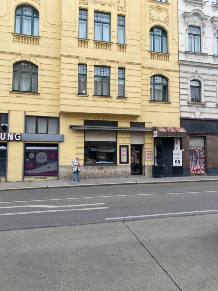 Straßenansicht eines gelben Altbaus mit dem Projektraum „Last Little Haven“ im Erdgeschoss. Das Schaufenster des Projektraums zeigt Plakate mit roter und weißer Gestaltung. Links vom Eingang steht eine Person mit heller Jacke und Mütze auf dem Gehsteig. Über dem Eingang hängt ein schwarzes Vordach, daneben die Hausnummer 68. Rechts daneben ist ein weiteres Geschäft mit dunklerer Fassade und Graffiti. Die Straße im Vordergrund hat Fahrbahnmarkierungen und einen breiten Gehsteig.