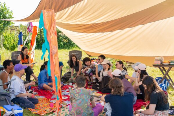 A group of people gather on a red cloth under a yellow/beige fabric canopy while conversing.