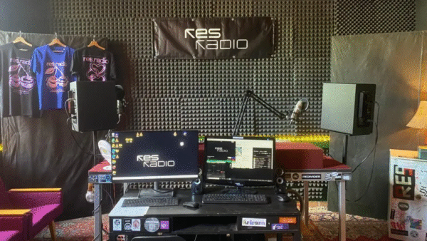 A picture of the res.radio studio. There is the DJ booth in the centre of it with three microphones, and there is a desk with two computers right in front of the booth. Behind - there is a big banner that says 