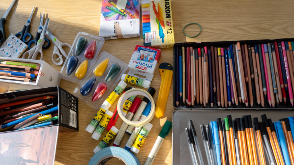 Bildbeschreibung: Aus der Vogelperspektive eine Nahaufnahme von vielen bunten Farben, Stifte, Wachskreiden, Scheren, Klebe- und Schneidutensilien auf einem sonnendurchfluteten Tisch versammelt. image description: A bird's-eye view close-up of many colourful pens, pencils, wax crayons, scissors, glue and cutting utensils gathered on a sun-drenched table.