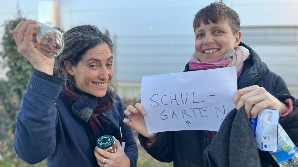 Zwei Personen stehen nebeinander in einem Garten und lächeln. Eine Person hält einen Zettel mit der Aufschrift 